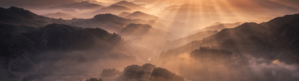 Sun rays shining over layered hills with morning mist under cloudy sky